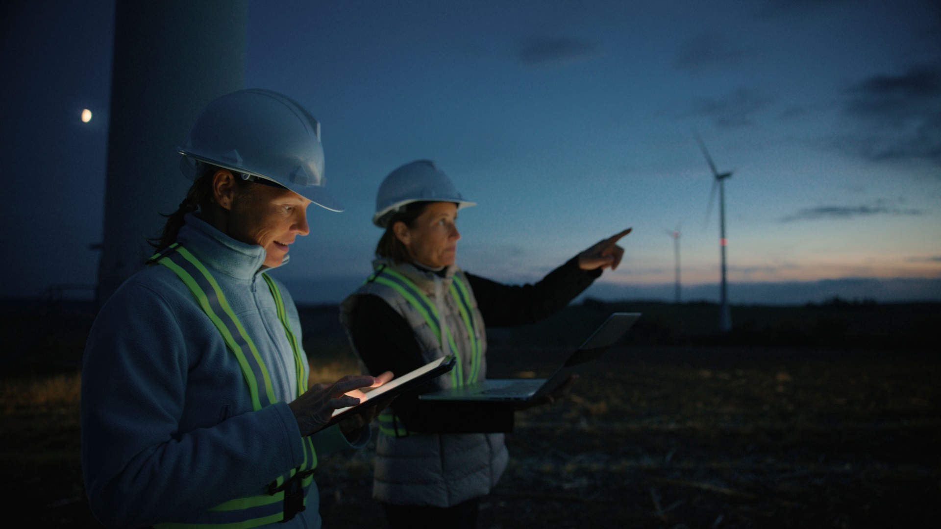 Female Engineers in Work Attire with Laptop and Digital Tablet Discussing About Wind Turbines on Farm at Sunset